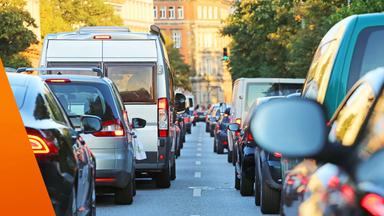 Der Blick führt zwischen zwei langen Reihen von Autos durch eine belebte Stadtstraße. Rote Rück- und Bremslichter prägen die Szene, in der Mitte steht ein großer weißer Lieferwagen. Die volle Straße veranschaulicht, dass viele Menschen trotz hoher Kraftstoffkosten weiterhin mit dem Auto unterwegs sind.