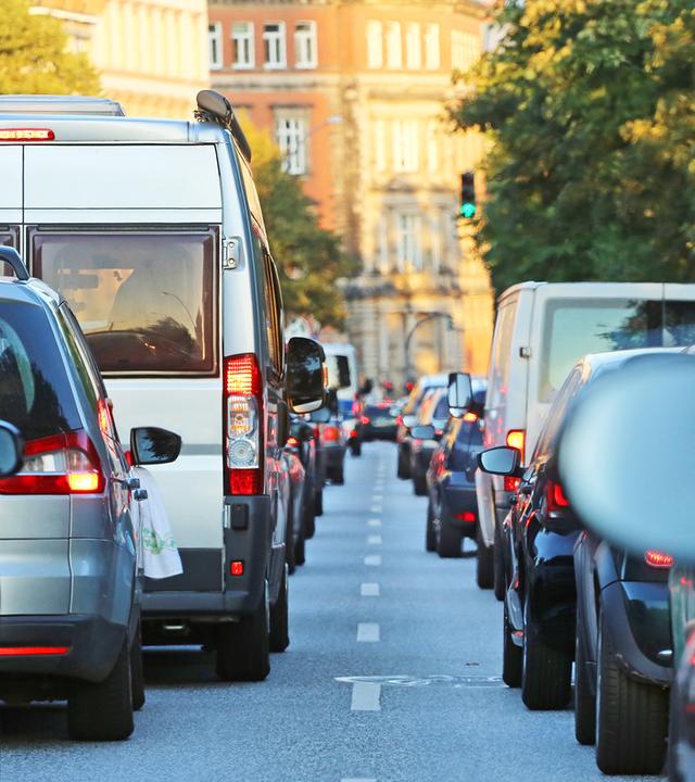 Der Blick führt zwischen zwei langen Reihen von Autos durch eine belebte Stadtstraße. Rote Rück- und Bremslichter prägen die Szene, in der Mitte steht ein großer weißer Lieferwagen. Die volle Straße veranschaulicht, dass viele Menschen trotz hoher Kraftstoffkosten weiterhin mit dem Auto unterwegs sind.