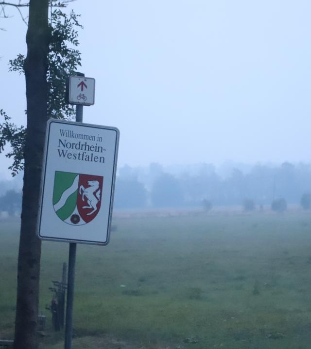 Rauch vom Waldbrand im Hohen Venn zieht über ein Feld, an dem ein Schild mit der Aufschrift "Willkommen in Nordrhein-Westfalen" die Staatsgrenze markiert.