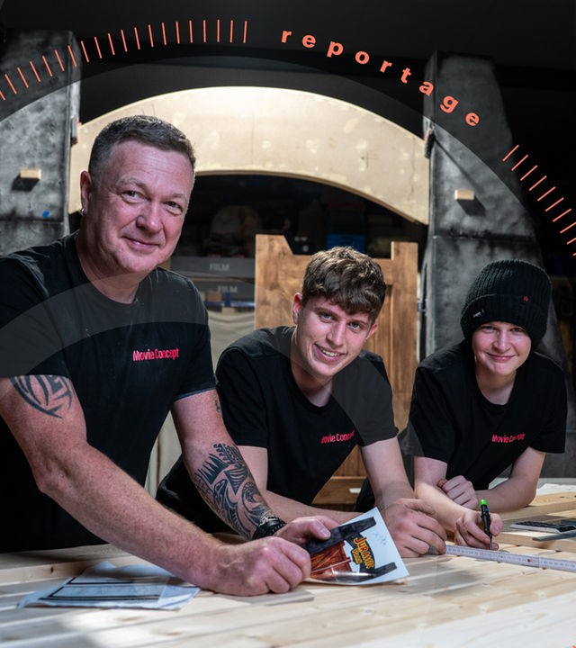Angelo Zannin und seine Söhne stehen an einer Tischlerplatte. Im Hintergrund steht schon ein Teil des Nachbaus des Eingangstors aus "Jurassic Park".