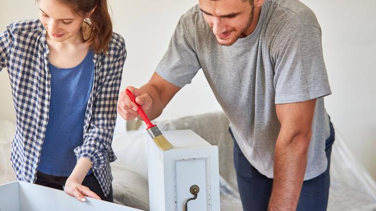 Ein Mann und eine Frau streichen die Schubladen eines Holzschranks weiß an.