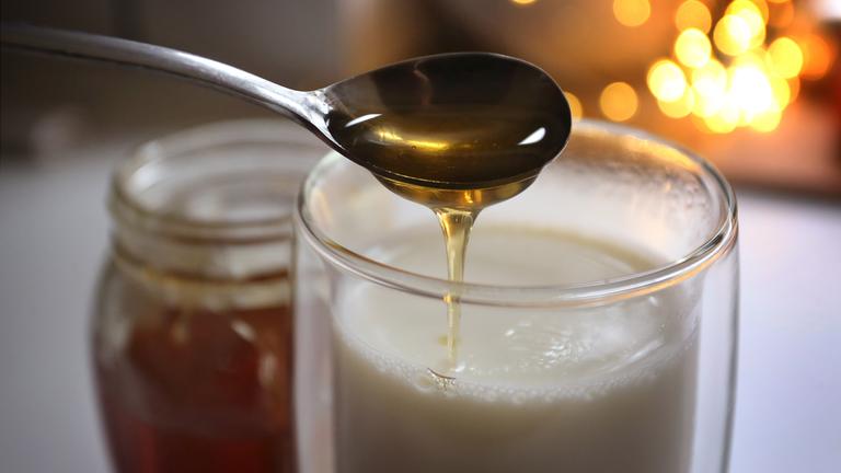 Es ist ein Glas heiße Milch mit einem Löffel Honig zu sehen.