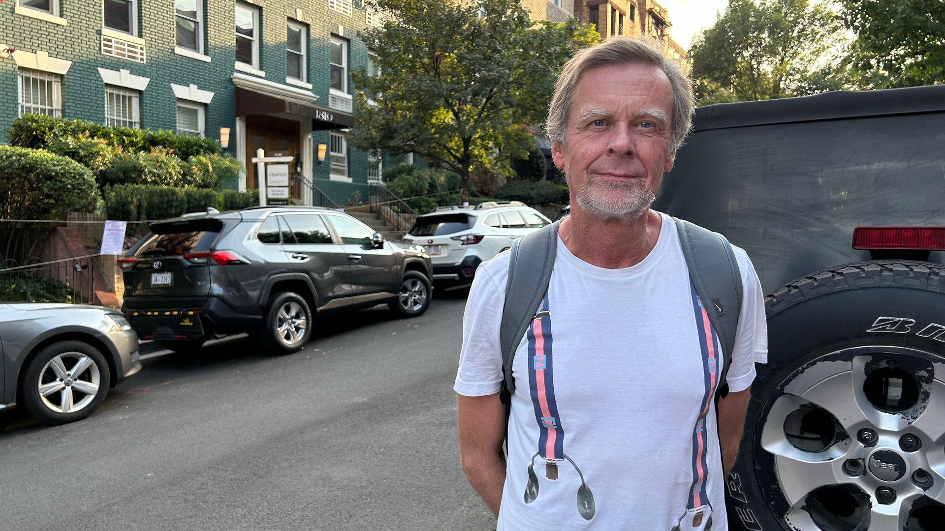 Ein Mann mit grauen Haaren steht im T-Shirt am Rand einer Straße.