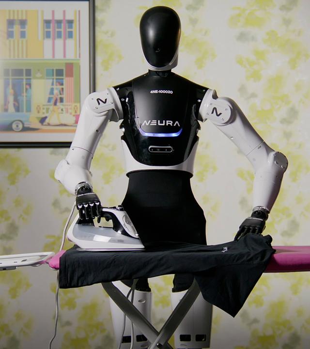 Ein schwarz-weißer humanoider Roboter steht vor einem Bügelbrett mit pinkfarbenem Bezug. Eine Hand liegt auf dem Griff eines Bügeleisens, die andere auf einem dunklen Kleidungsstück, das über das Brett ausgebreitet ist. Auf der Brust des Roboters steht „NEURA“. Im Hintergrund sind eine geblümte Tapete, ein gerahmtes Bild und eine Stehlampe zu sehen.