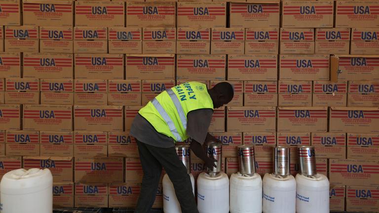 Ein Mann in Warnweste verräumt Zylinder vor einer Wand aus Kartons.