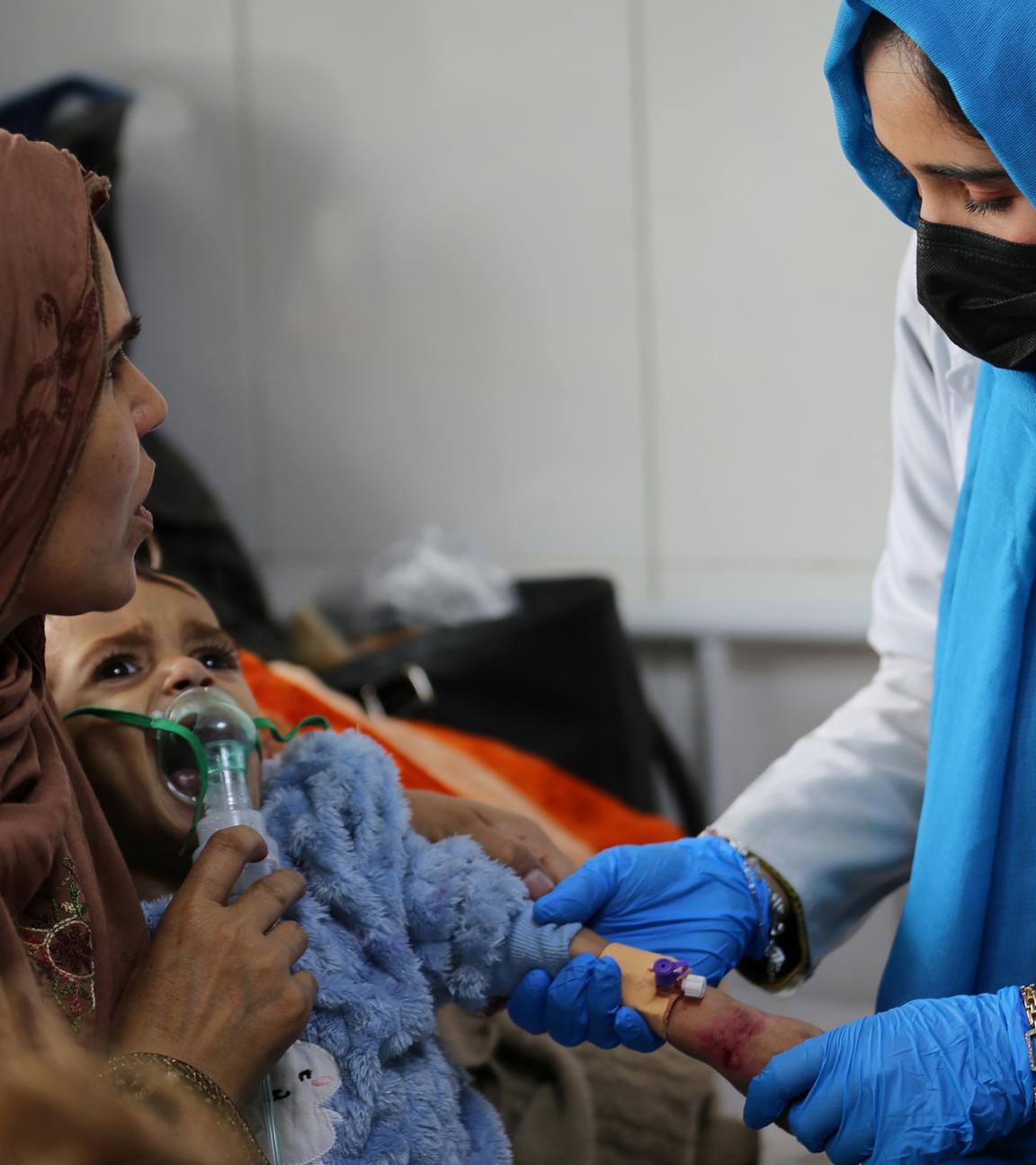 Latifa Bakkar versucht, ihrem schwer unterernährten 2½-jährigen Sohn Abu Bakar Sauerstoff zu verabreichen, während eine Krankenschwester ihm im Indira-Gandhi-Kinderkrankenhaus in Kabul, Afghanistan, am  15. 2. 2026, eine intravenöse Kanüle in den Arm legt. 