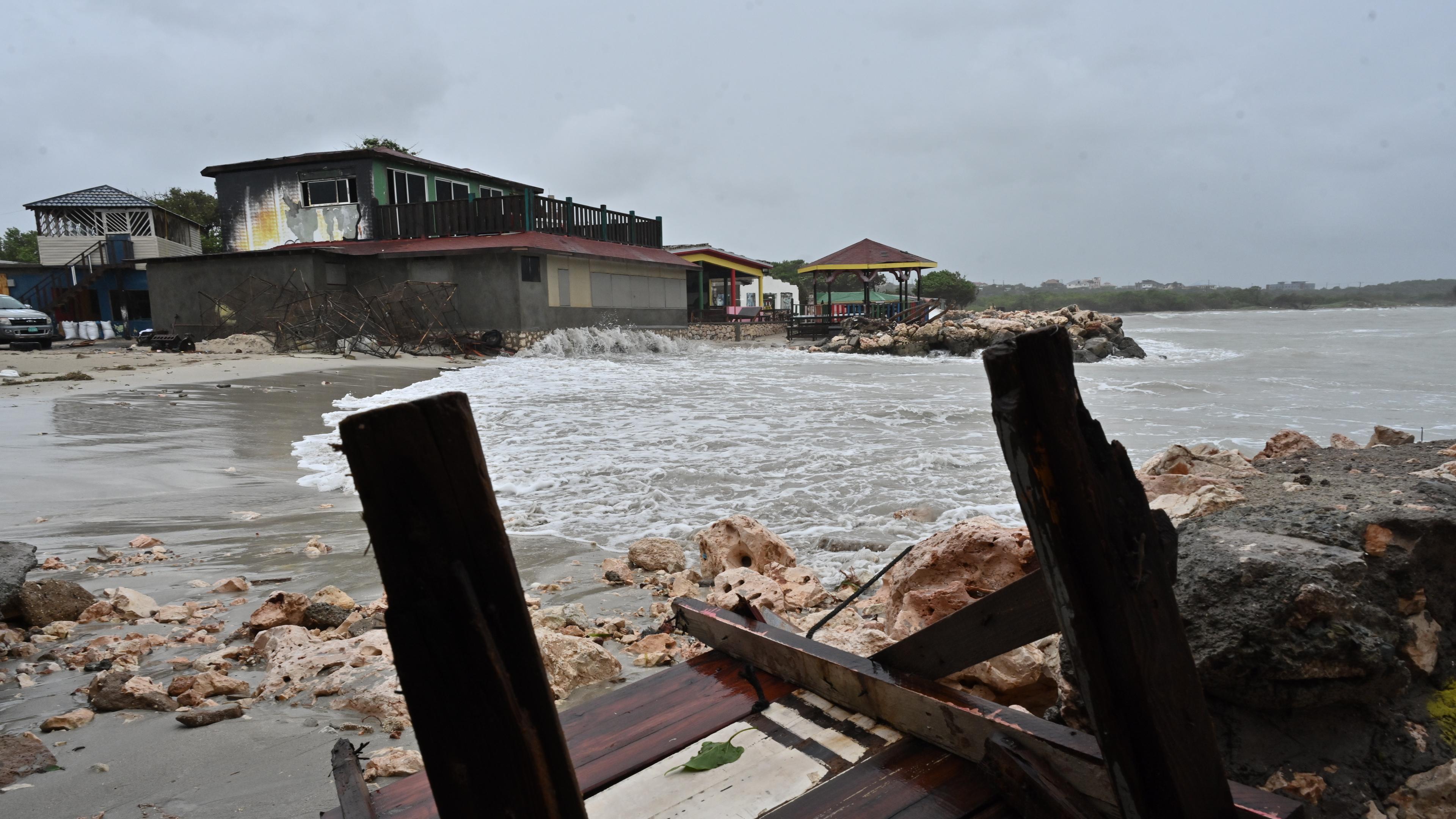 Es ist eine Küste Jamaikas zu sehen. Holzmöbel liegen zertört am Strand und der Himmel ist düster.