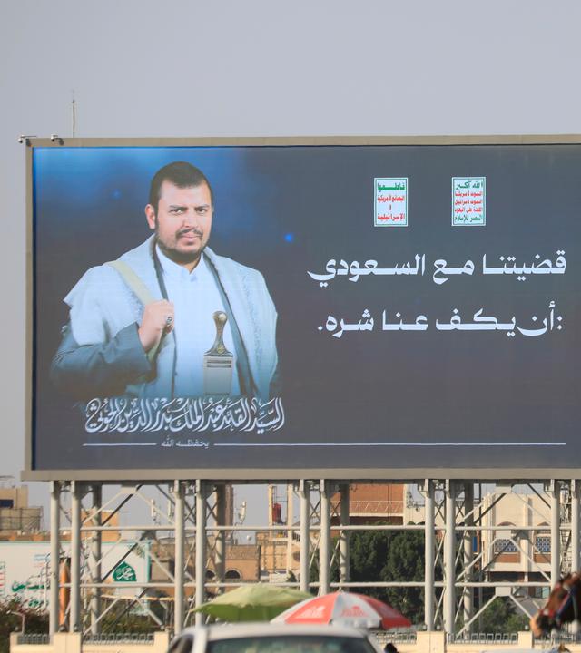 Jemenitische Flaggen wehen neben einer digitalen Werbetafel, die den Huthi-Anführer Abdul-Malik al-Huthi sowie den arabischen Schriftzug „Unser Streit mit Saudi-Arabien besteht darin, dass es seine Aggressionen gegen uns einstellt“ zeigt.