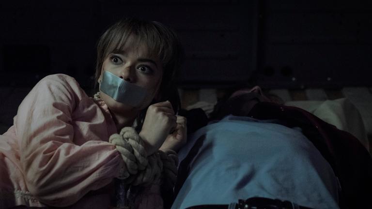 Eine Frau mit verbundenen Händen und Klebeband über dem Mund, neben ihr liegt ein Mann auf dem Rücken.