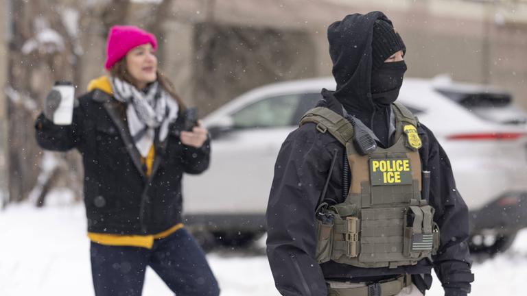 Ein Polizist und im Hintergrund unscharf eine Frau während eines ICE-Einsatzes nahe der Karmel Mall am 10.12.2025 in Minneapolis, USA.