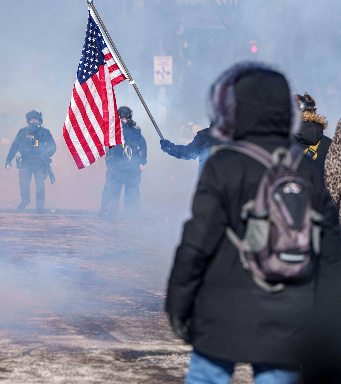 Mit dem Rücken zum Betrachter stehen demonstrierende Menschen mit einer US-Flagge bewaffneten Mitgliedern der ICE-Beamten gegenüber.