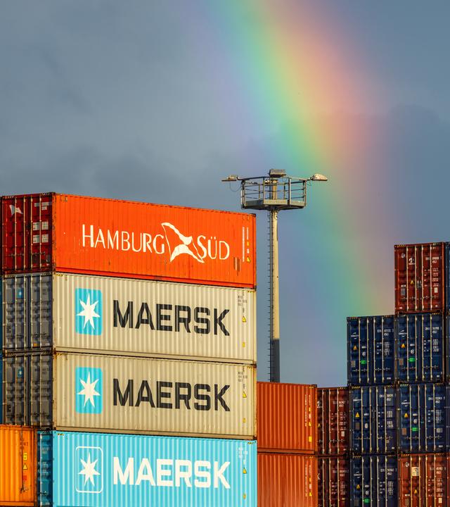 Symbolbild: Gestapelte Frachtcontainer der Reedereien Maersk, Hamburg Sued und Hapag-Lloyd auf einem Containerterminal. Ein Regenbogen über dem Hafengelände markiert einen Wetterwechsel.