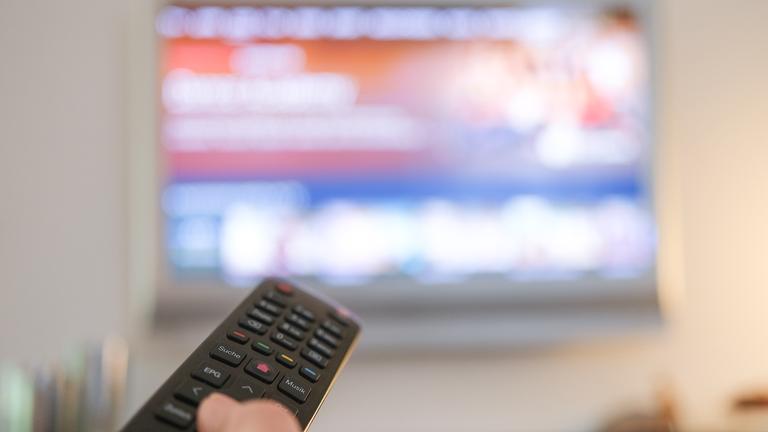 Eine Person hält eine Fernbedienung in Richtung Fernseher, welcher leicht verschwommen im Hintergrund zu sehen ist.