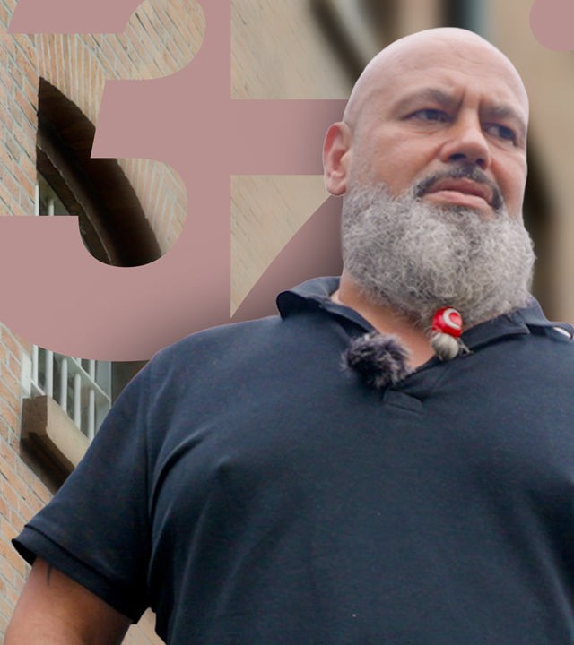 Untersicht: Berkmann ist auf dem Außengelände der JVA. Er steht auf einem betonierten Platz, der von hohen Metallzäunen umgeben ist. Er schaut seitlich in die Ferne.