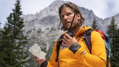 Bergretter Markus Kofler (Sebastian Ströbel) steht auf einer Wiese zwischen Bäumen und spricht in ein Funkgerät. In der anderen Hand hält er einen abgerissenen Zettel. Im Hintergrund Berge.