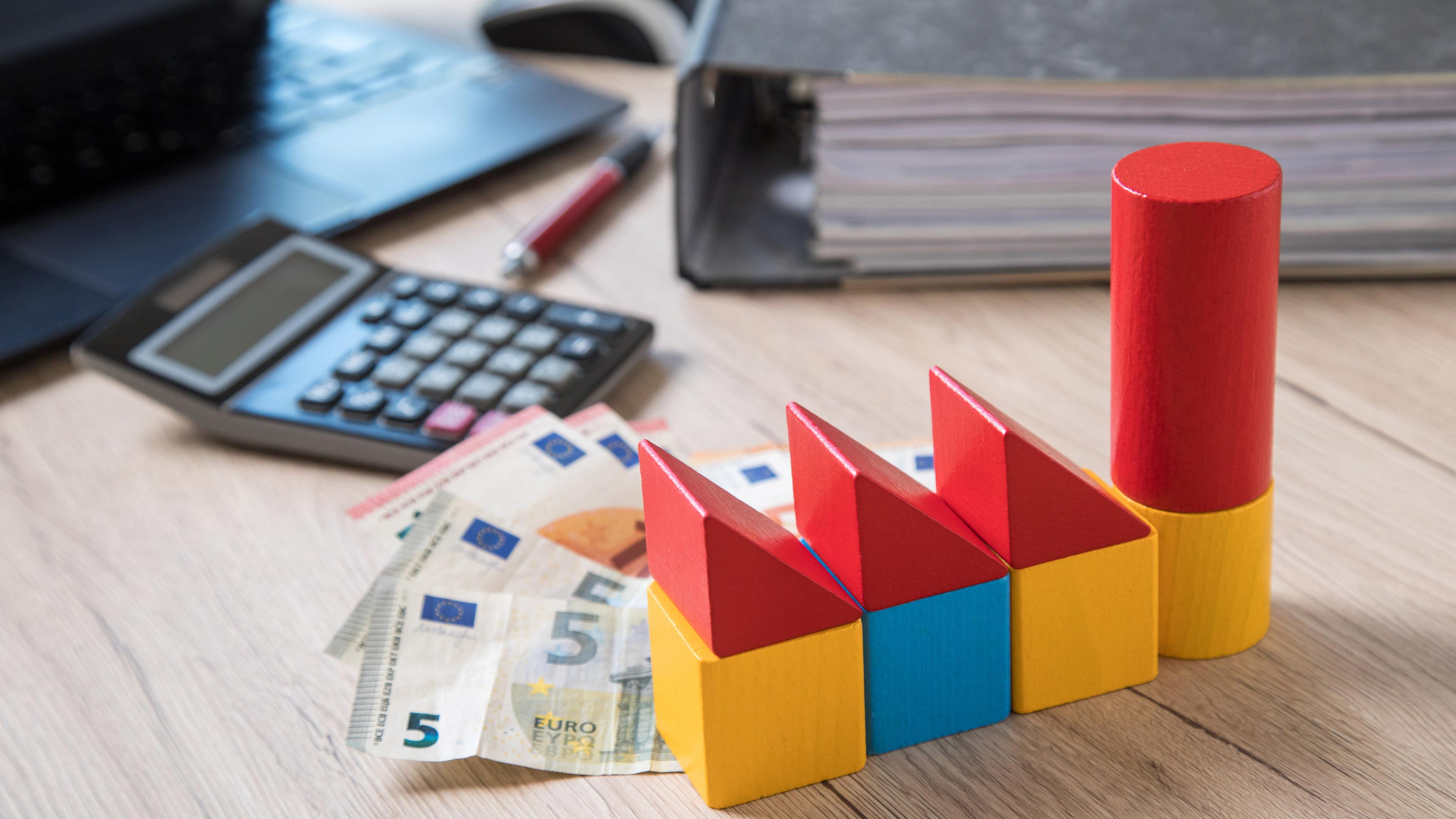 Bauklötze, Bargeld, ein Taschenrechner, ein Aktenordner und ein Laptop auf einem Tisch
