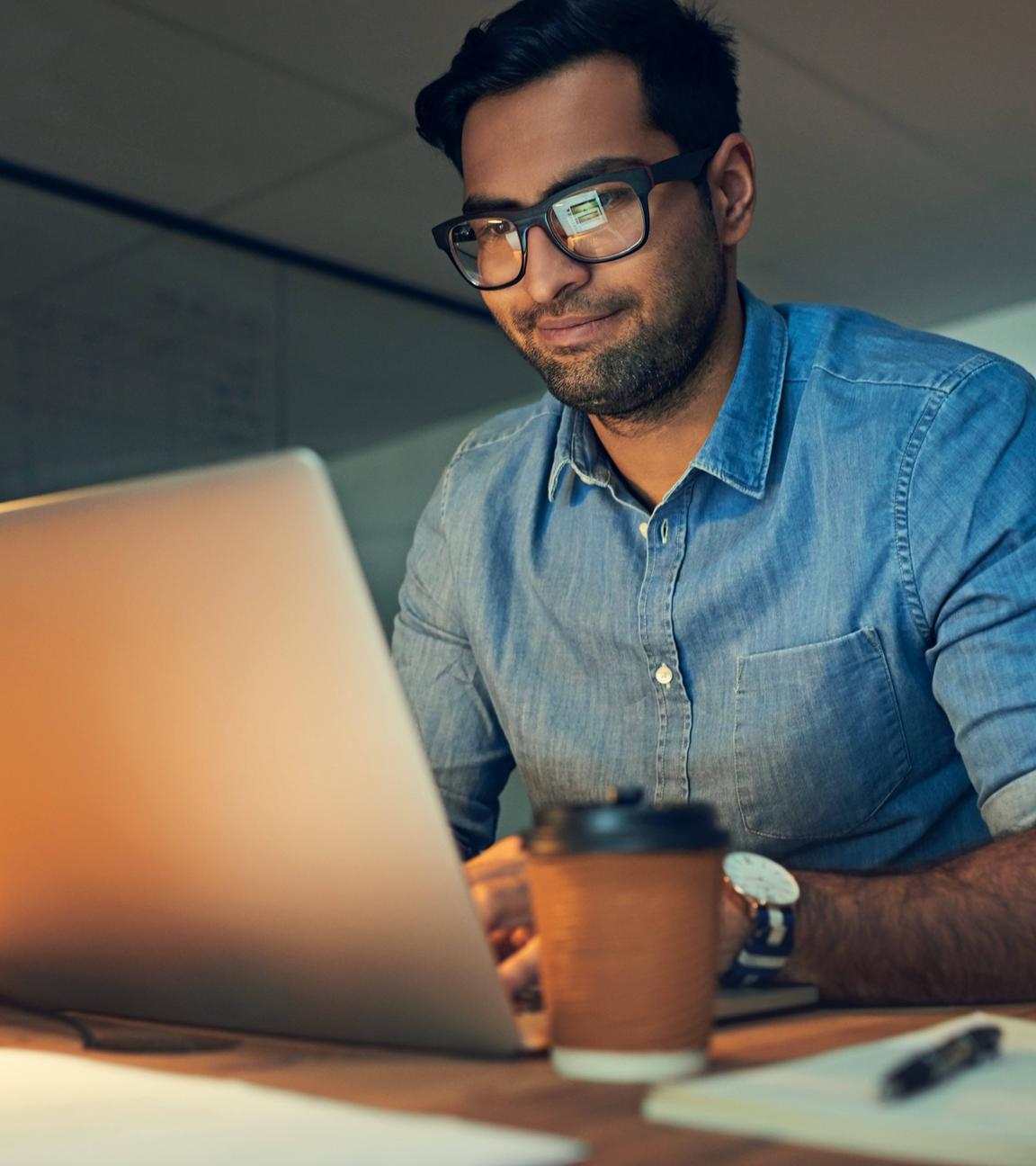 Ein indischer Mann sitzt nachts an einem beleuchteten Schreibtisch mit einem Laptop vor sich.