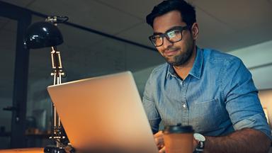 Ein indischer Mann sitzt nachts an einem beleuchteten Schreibtisch mit einem Laptop vor sich.
