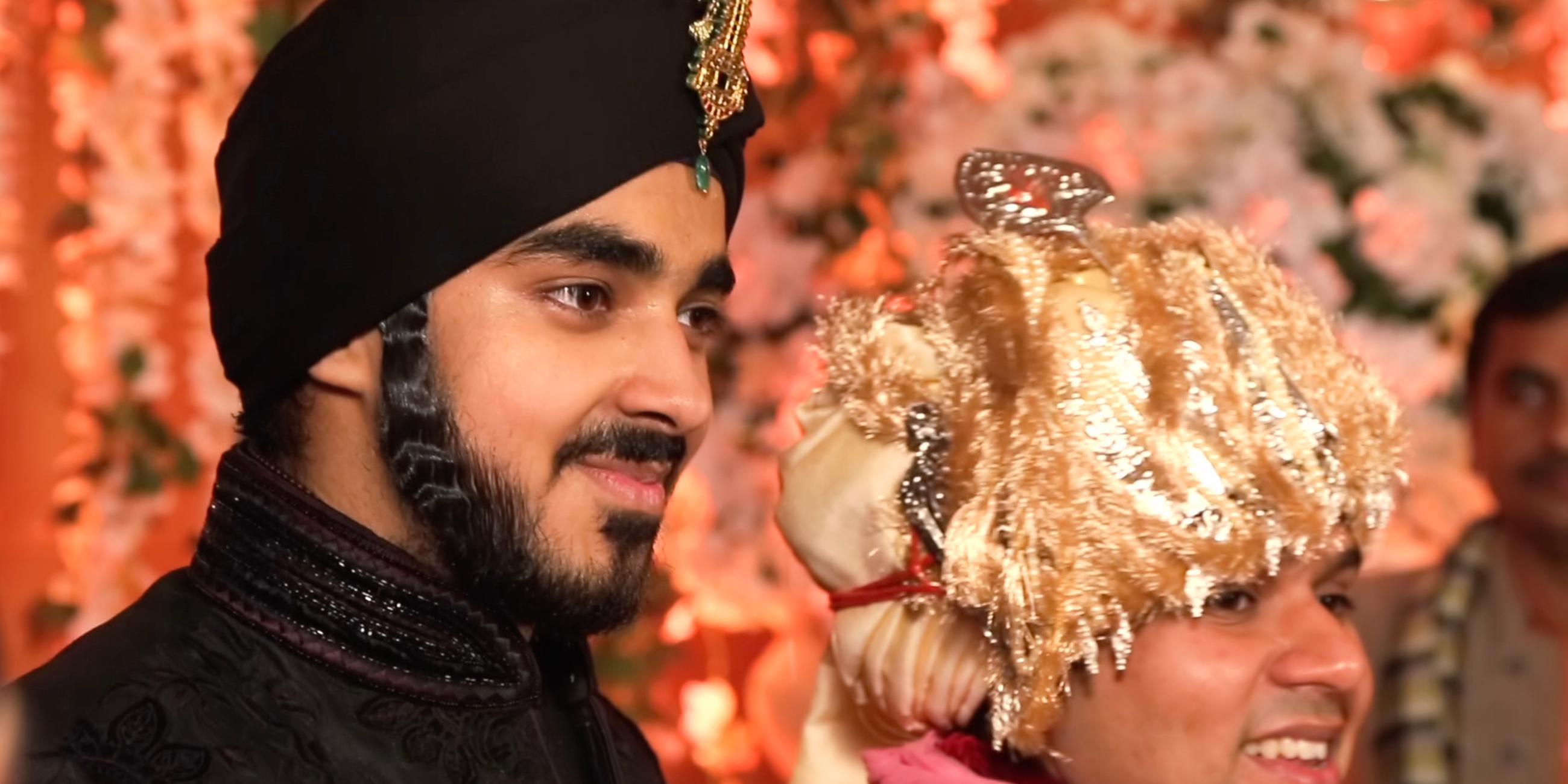 "Indiens Superreiche - Zwischen Elend und Luxus": Evan Luthra steht neben einem jungen Mann. Luthra trägt eine edle schwarze und hochgeschlossene Robe mit schwarzem Turban, den mittig über der Stirn ein goldenes Schmuckstück ziert. Der Mann neben ihm trägt einen aufwändigen goldenen Kopfschmuck. Beide lächeln.