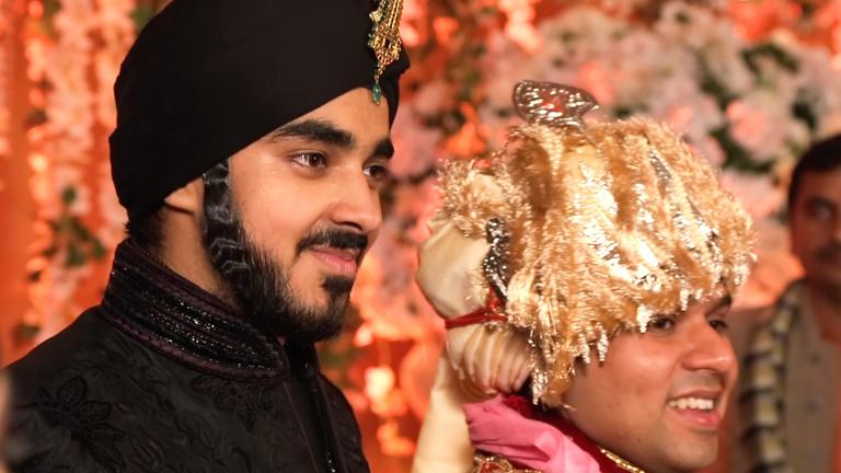 "Indiens Superreiche - Zwischen Elend und Luxus": Evan Luthra steht neben einem jungen Mann. Luthra trägt eine edle schwarze und hochgeschlossene Robe mit schwarzem Turban, den mittig über der Stirn ein goldenes Schmuckstück ziert. Der Mann neben ihm trägt einen aufwändigen goldenen Kopfschmuck. Beide lächeln.