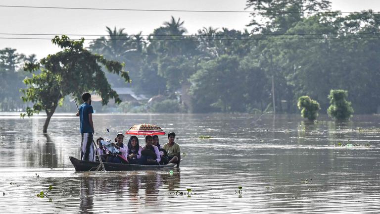 Ein Boot/Floß mit Menschen treibt auf überfluteter Fläche, im Hintergrund ist ein Wald