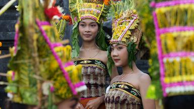 Zwei junge asiatisch aussehende Frauen tragen traditionellen balinesischen Kopfschmuck aus Blumen und Blättern und bestickte schulterfreie Wickelkleider.