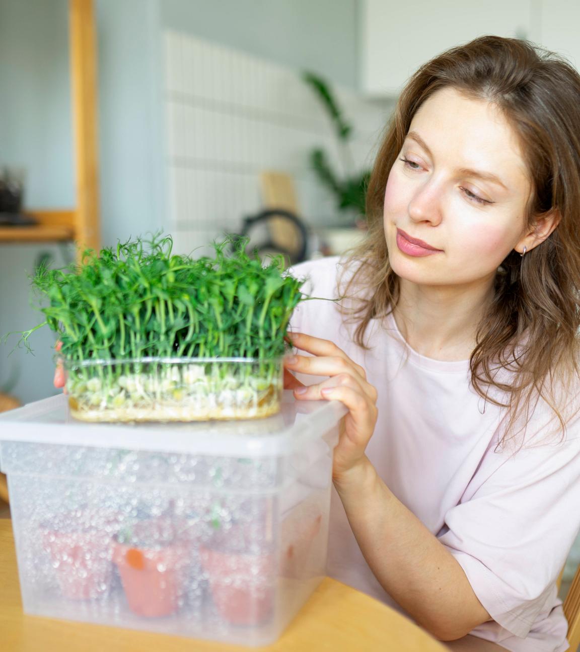 Eine Frau sitzt am Küchentisch, vor ihr eine Plastikbox mit kleinen Pflanztöpfen, auf der eine Schale mit frischer Kresse steht.
