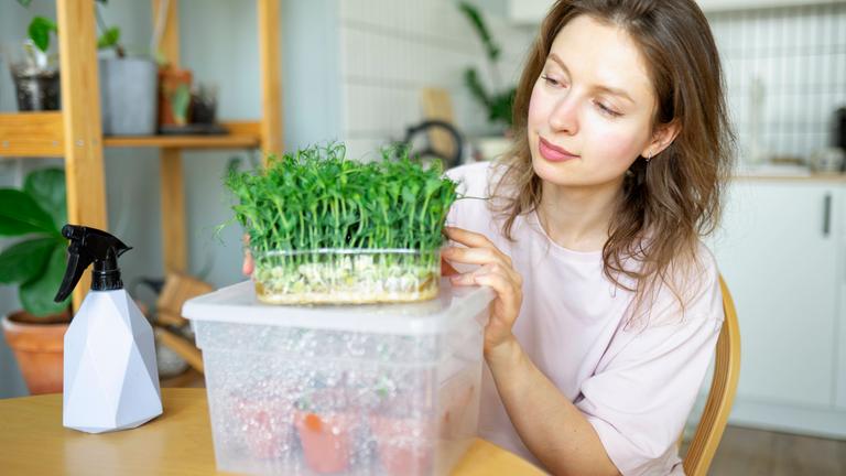 Eine Frau sitzt am Küchentisch, vor ihr eine Plastikbox mit kleinen Pflanztöpfen, auf der eine Schale mit frischer Kresse steht.