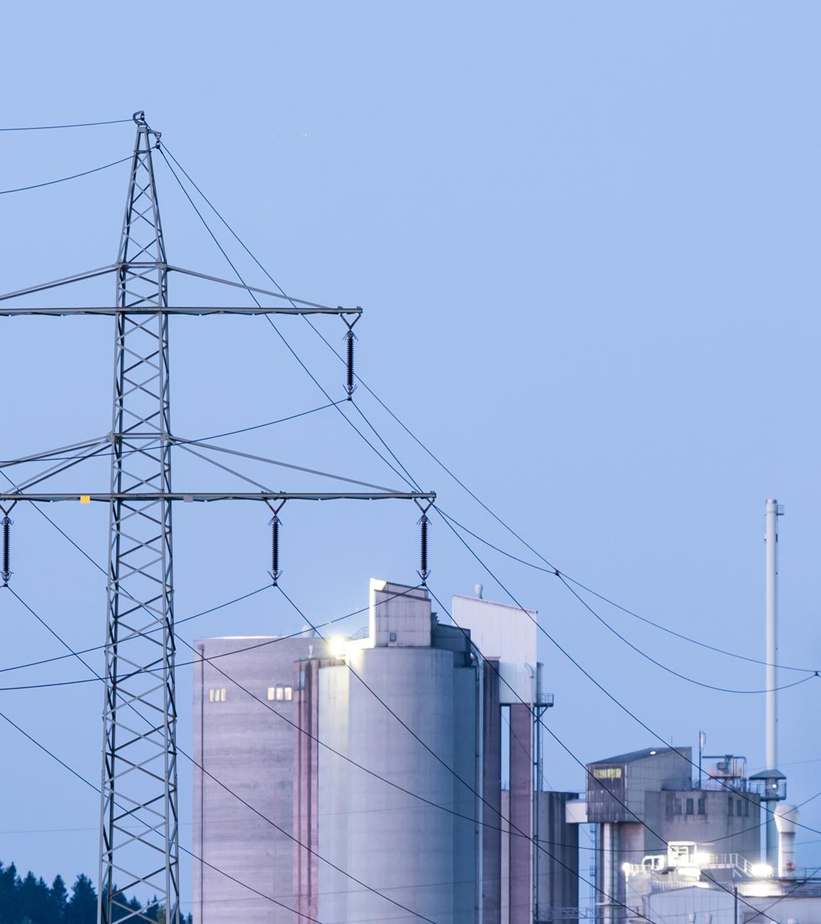 Strommasten stehen im Morgenlicht vor einem Zementwerk. 