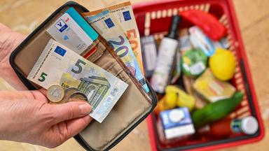 Eine Frau hält auf ihrer Hand Geld vor einem vollen Einkaufskorb mit Lebensmitteln.