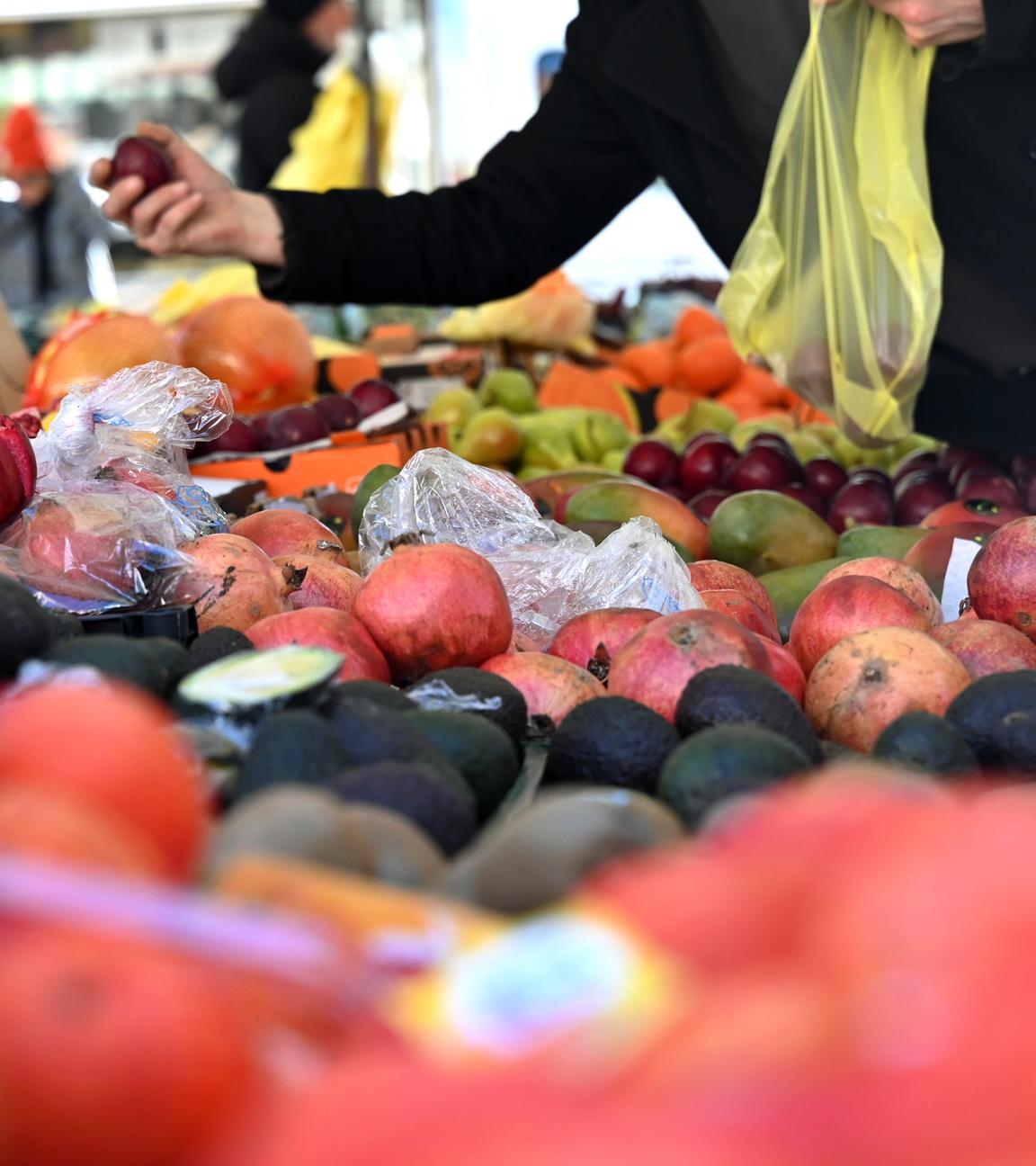 Eine Person hält an einem Obststand am Marktplatz in Leipzig eine Pflaume und eine Einkaufstüte in der Hand.