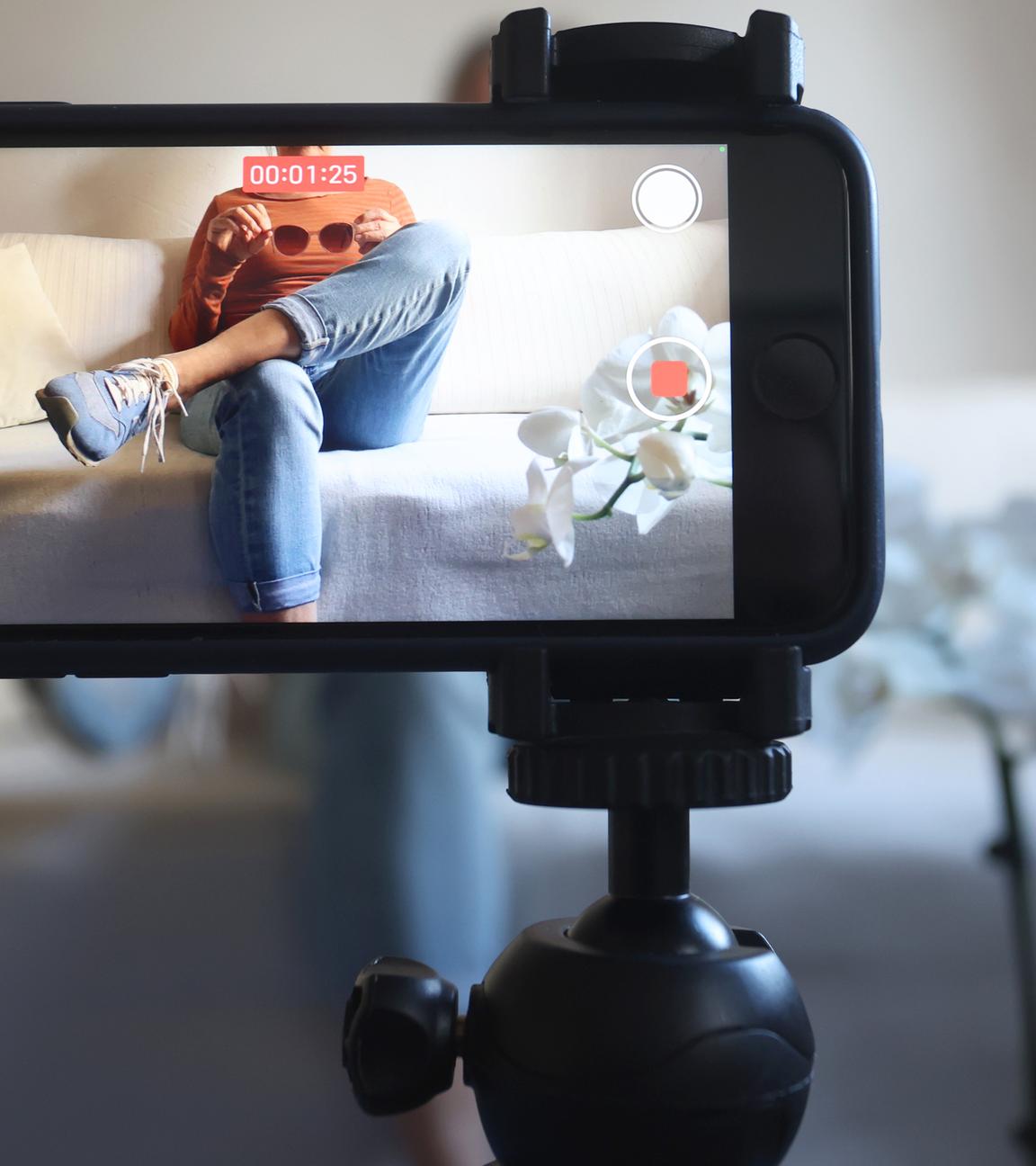 Eine Frau sitzt mit einer Sonnenbrille in Händen auf einem Sofa und filmt sich dabei mit ihrem iPhone.