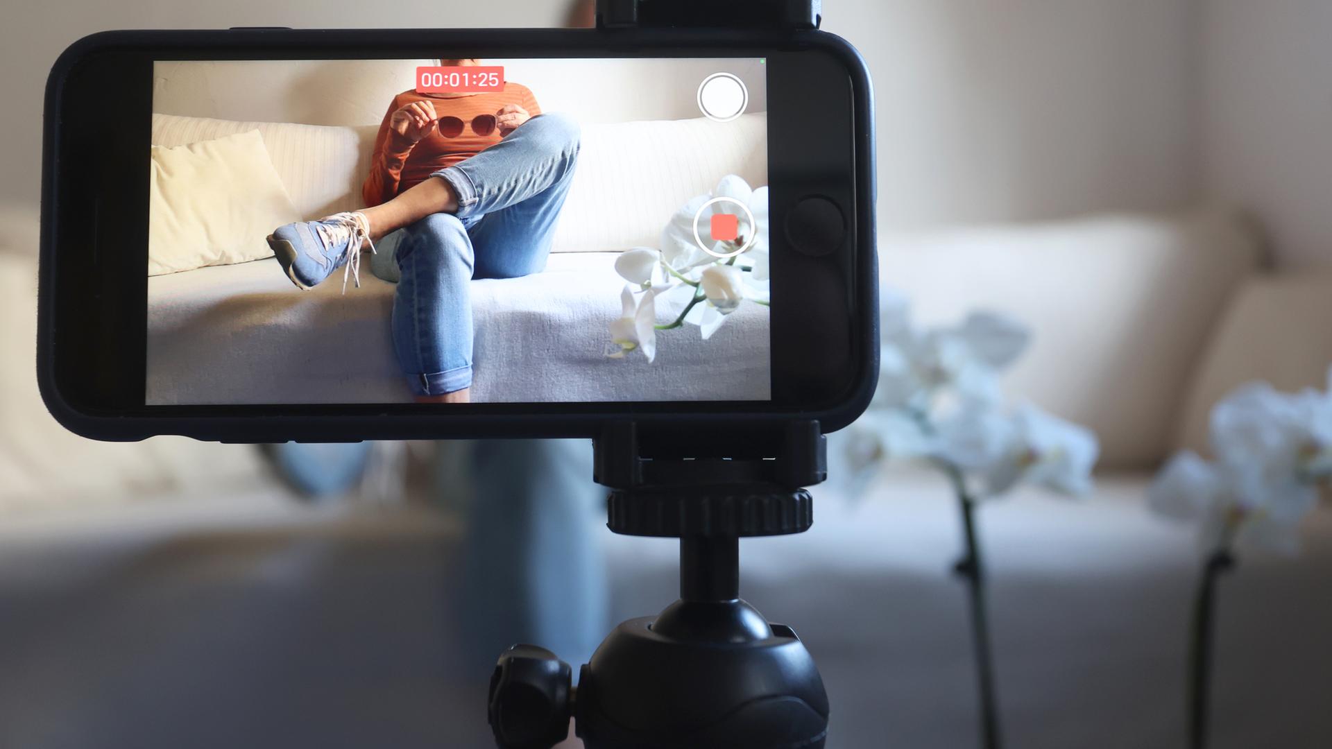 Eine Frau sitzt mit einer Sonnenbrille in Händen auf einem Sofa und filmt sich dabei mit ihrem iPhone.