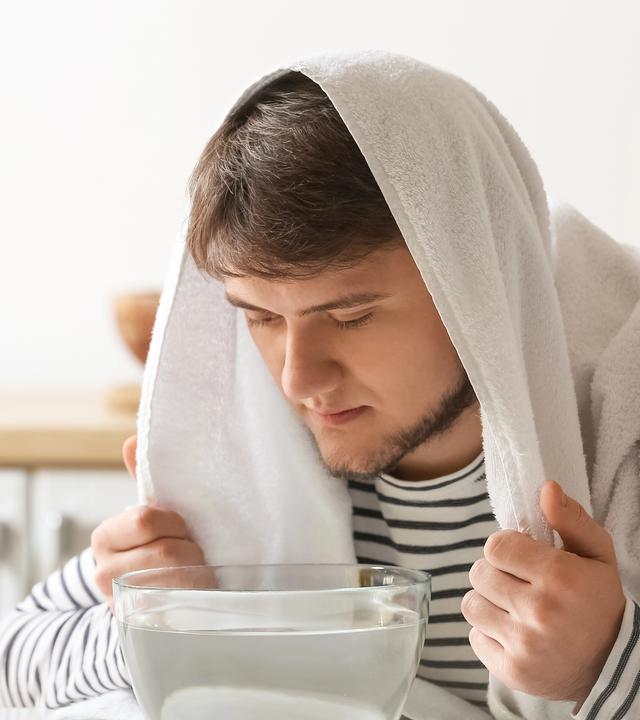 Ein Mann mit einem Handtuch über dem Kopf sitzt vor einer Schale mit heißem Wasser in einer Küche.