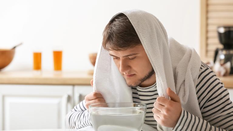 Ein Mann mit einem Handtuch über dem Kopf sitzt vor einer Schale mit heißem Wasser in einer Küche.
