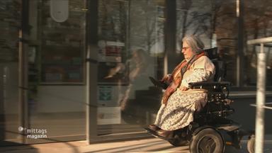 Frau im Rollstuhl fährt eigenständig über eine Straße, sie lenkt ihren Rollstuhl über einen Schalter mit der Hand