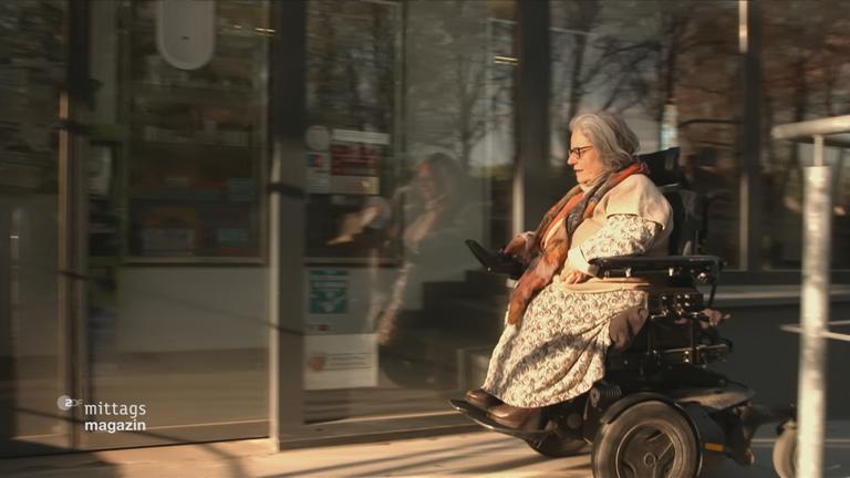 Frau im Rollstuhl fährt eigenständig über eine Straße, sie lenkt ihren Rollstuhl über einen Schalter mit der Hand