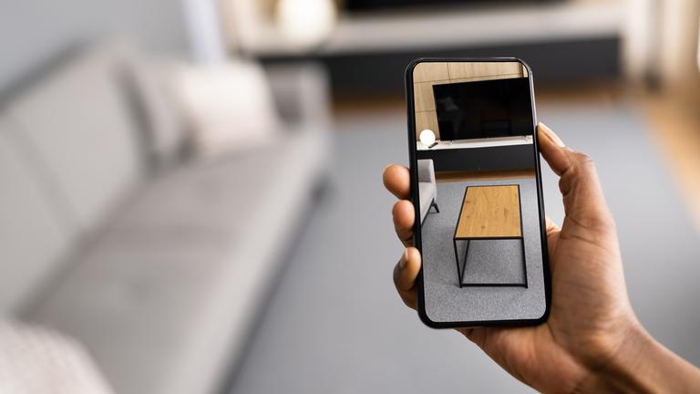 Ein Männ hält ein Smartphone, auf dessen Bildschirm ein Couchtisch vor einem Sofa dargestellt wird, in leeres Wohnzimmer.