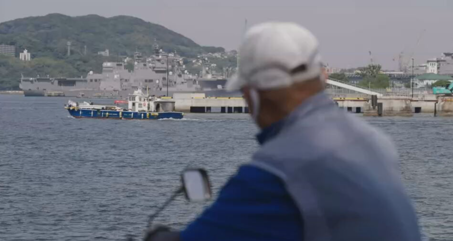 Im Vordergrund der Rücken eines Mannes, der auf einen Hafen blickt. Marineschiff im Hintergrund.