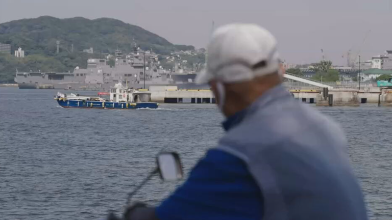 Im Vordergrund der Rücken eines Mannes, der auf einen Hafen blickt. Marineschiff im Hintergrund.