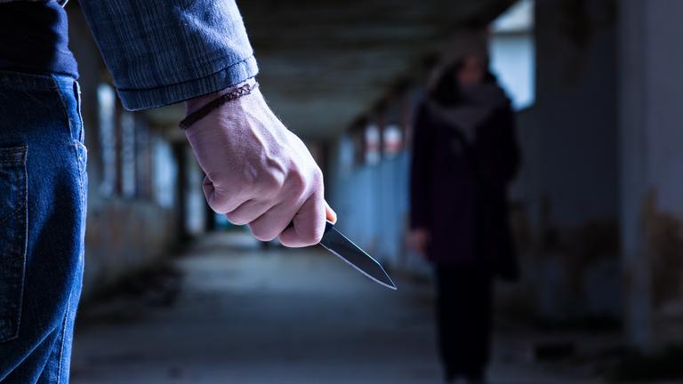 "Inside the Criminal Mind: Serienkiller": Im Vordergrund ist eine männliche Hand zu sehen, die ein Messer hält. Eine Frau ist verschwommen im Hintergrund zu erkennen.