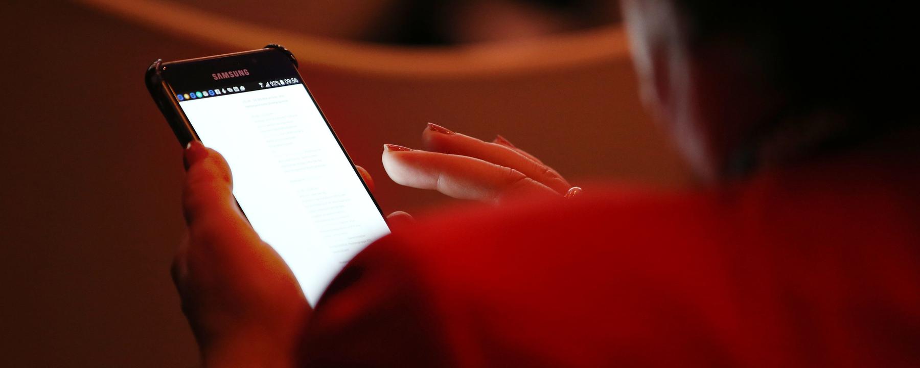 Eine Person ist von hinten in einem dunklen Raum zu sehen und hält ihr weiß leuchtendes Smartphone in der Hand.