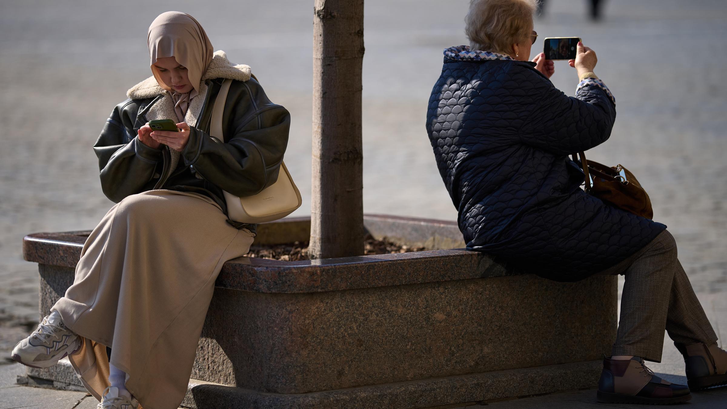 Eine Frau mit Kopftuch und eine weitere Frau sitzen am Roten Platz in Moskau und halten jeweils ein Smartphone in der Hand.