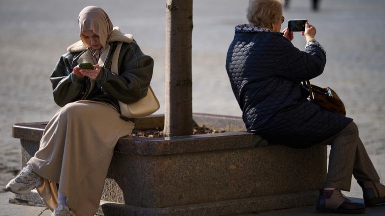 Eine Frau mit Kopftuch und eine weitere Frau sitzen am Roten Platz in Moskau und halten jeweils ein Smartphone in der Hand.