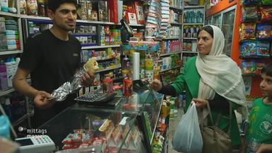 Auf dem Bild befinden sich drei Leute in einem kleinen Supermarkt. Auf der einen Seite des Tresens bei der Kasse steht ein Mann im schwarzen T-shirt und auf der anderen Seite eine Frau und neben ihr ein kleiner Junge. Die Frau hält eine Einkaufstüte in der Hand.