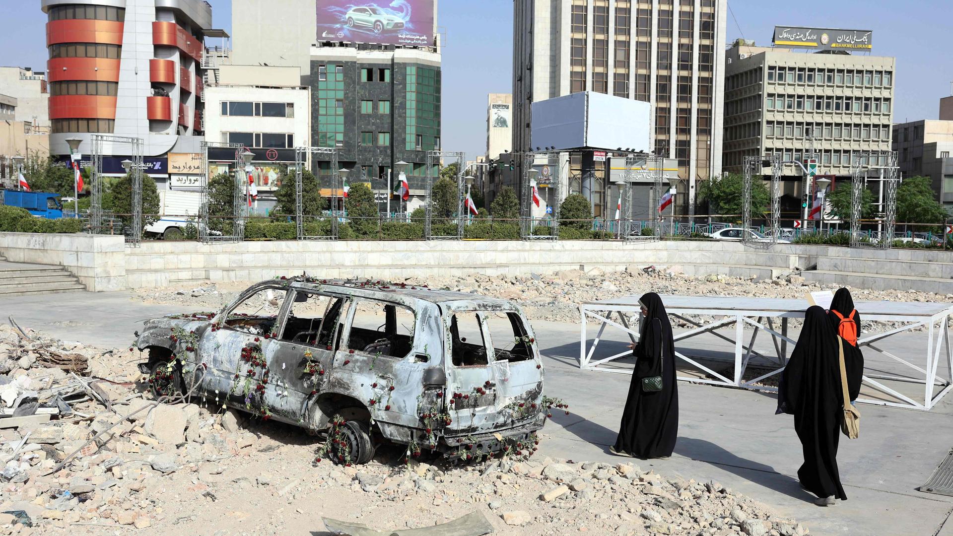 Ein zerstörtes Fahrzeug auf einer zerstörten Fläche in Iran, drei Menschen in schwarzen Gewändern stehen daneben.