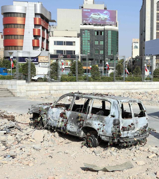 Ein zerstörtes Fahrzeug auf einer zerstörten Fläche in Iran, drei Menschen in schwarzen Gewändern stehen daneben.