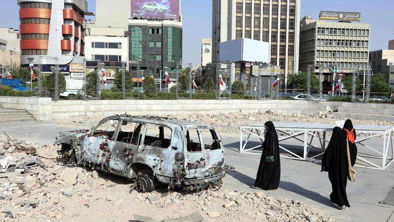 Ein zerstörtes Fahrzeug auf einer zerstörten Fläche in Iran, drei Menschen in schwarzen Gewändern stehen daneben.