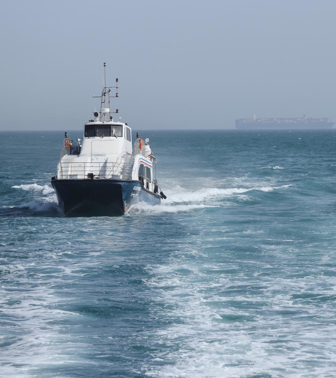 Iran, Bandar Abbas: Ein Patrouillenboot fährt durch das Wasser, während Frachtschiffe in der Straße von Hormuz vor Bandar Abbas, Iran, vor Anker liegen.