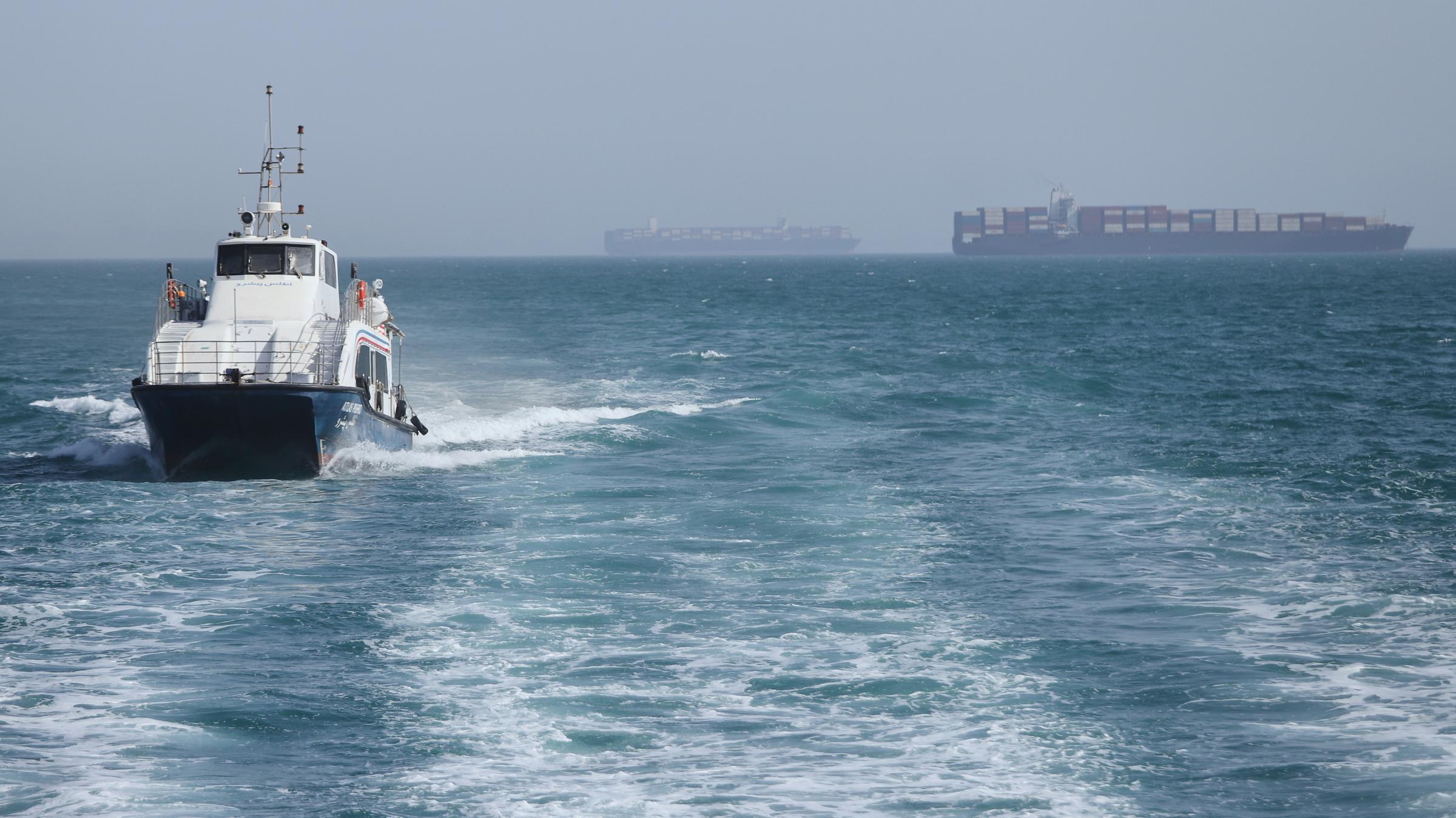 Iran, Bandar Abbas: Ein Patrouillenboot fährt durch das Wasser, während Frachtschiffe in der Straße von Hormuz vor Bandar Abbas, Iran, vor Anker liegen.